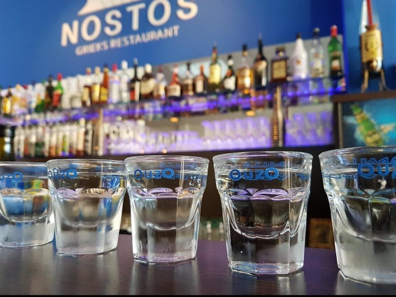Glaasjes ouzo op de bar van Grieks Restaurant Nostos, met het blauwe Nostos-logo op de achtergrond